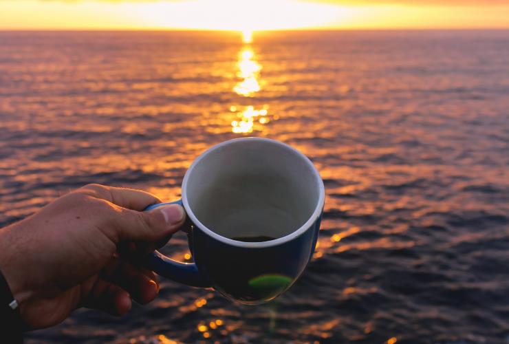 A person holding a blue coffee cup while looking at the sunrise over the sea.